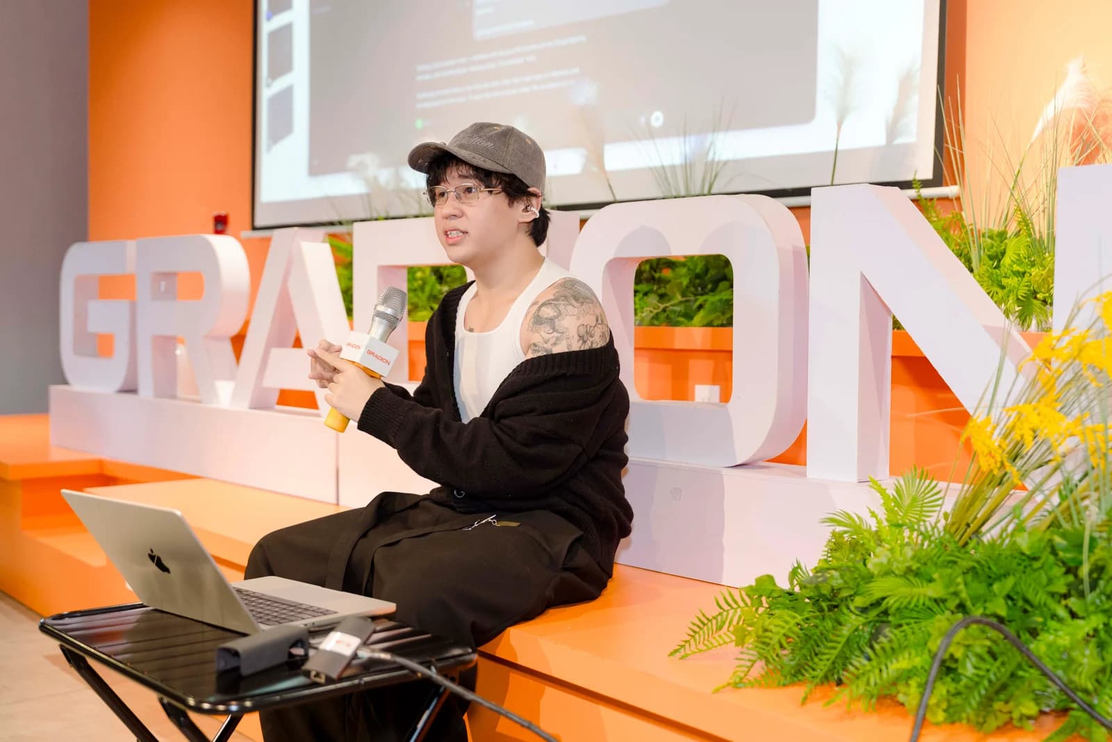 A speaker seated on the edge of the stage with a microphone and an open laptop nearby.